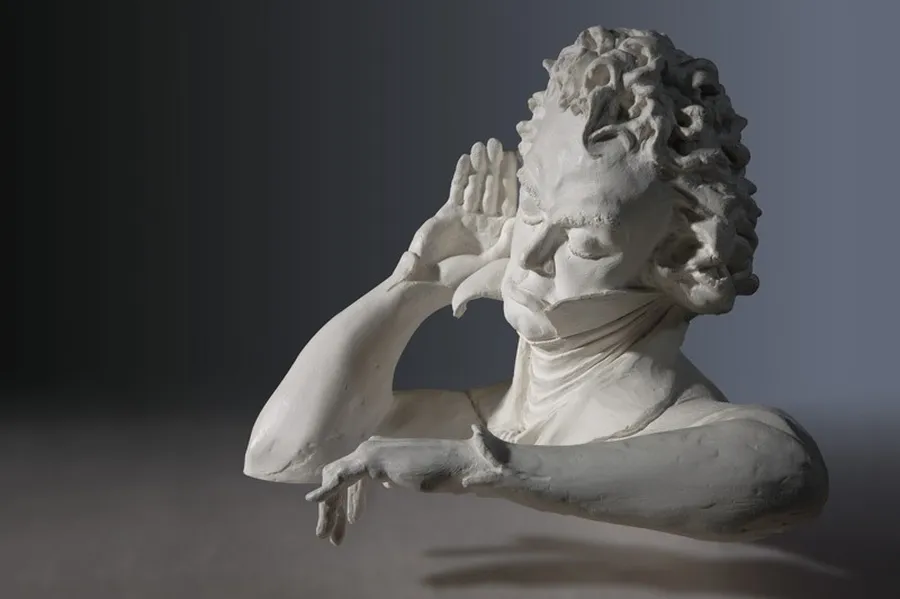 Beethoven holds his hand to his ear to hear better, as a modern white plaster bust shows him.
