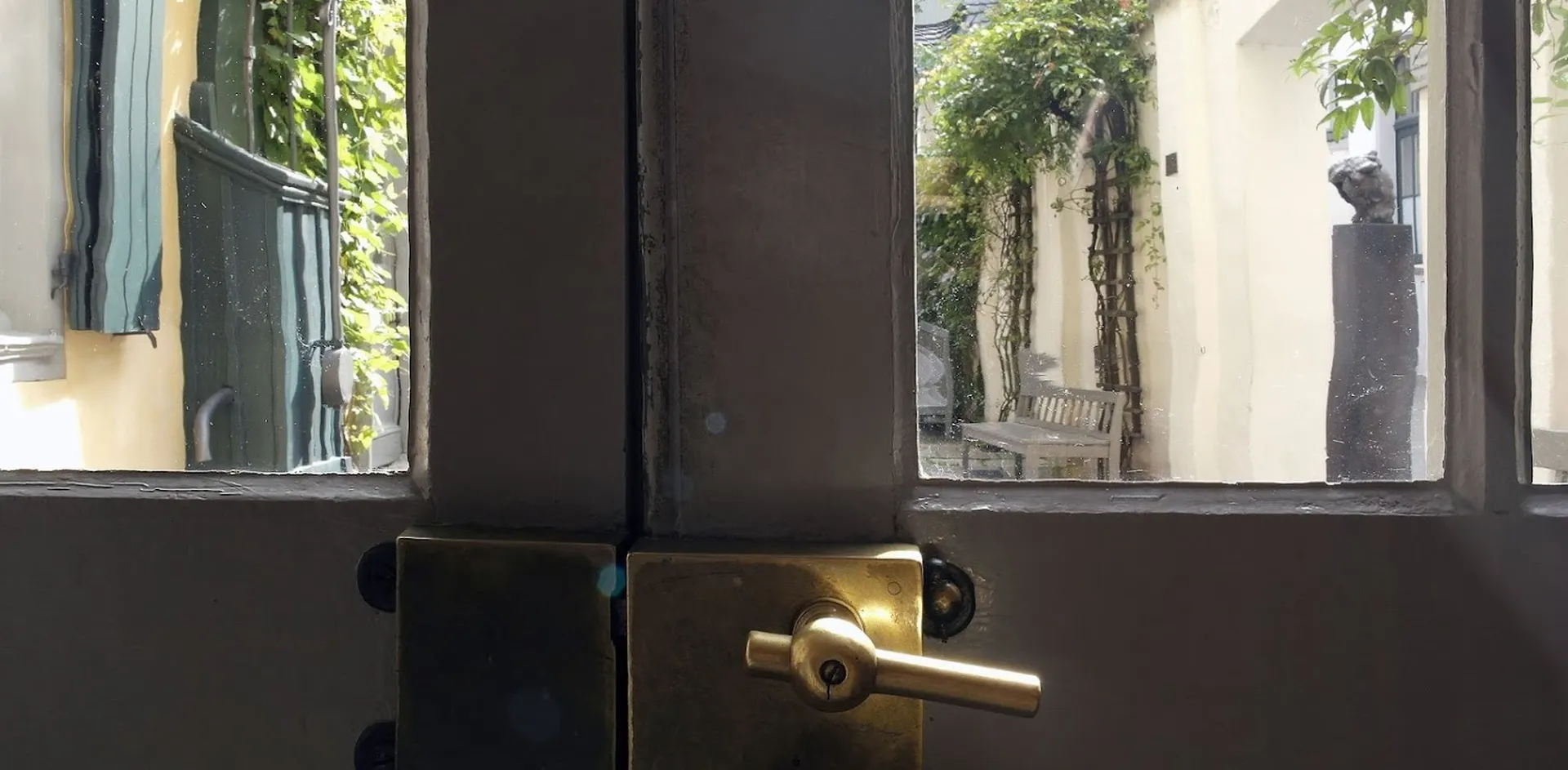 Ein Lichtstrahl fällt auf den Messingbeschlag einer Tür zum Museumshof. Durch die beiden Türfenster sieht man links eine Teil der Fassade von Beethovens Geburts- und Wohnhaus und rechts eine Holzbank vor einer Mauer mit Kletterpflanzen.